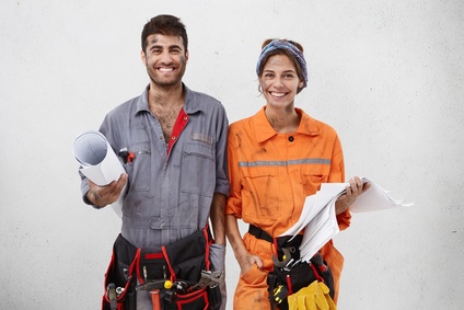 Joyful handyman and female architect collaborate together, wear working clothes, hold sketches with new construction plan, smile broadly, stand next to each other over white blank studio wall